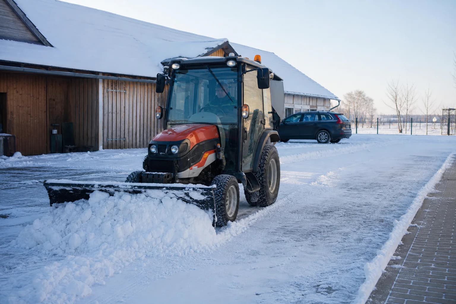 Malý traktor s kabínou a prednou radlicou odhŕňa sneh na hospodárskom dvore.