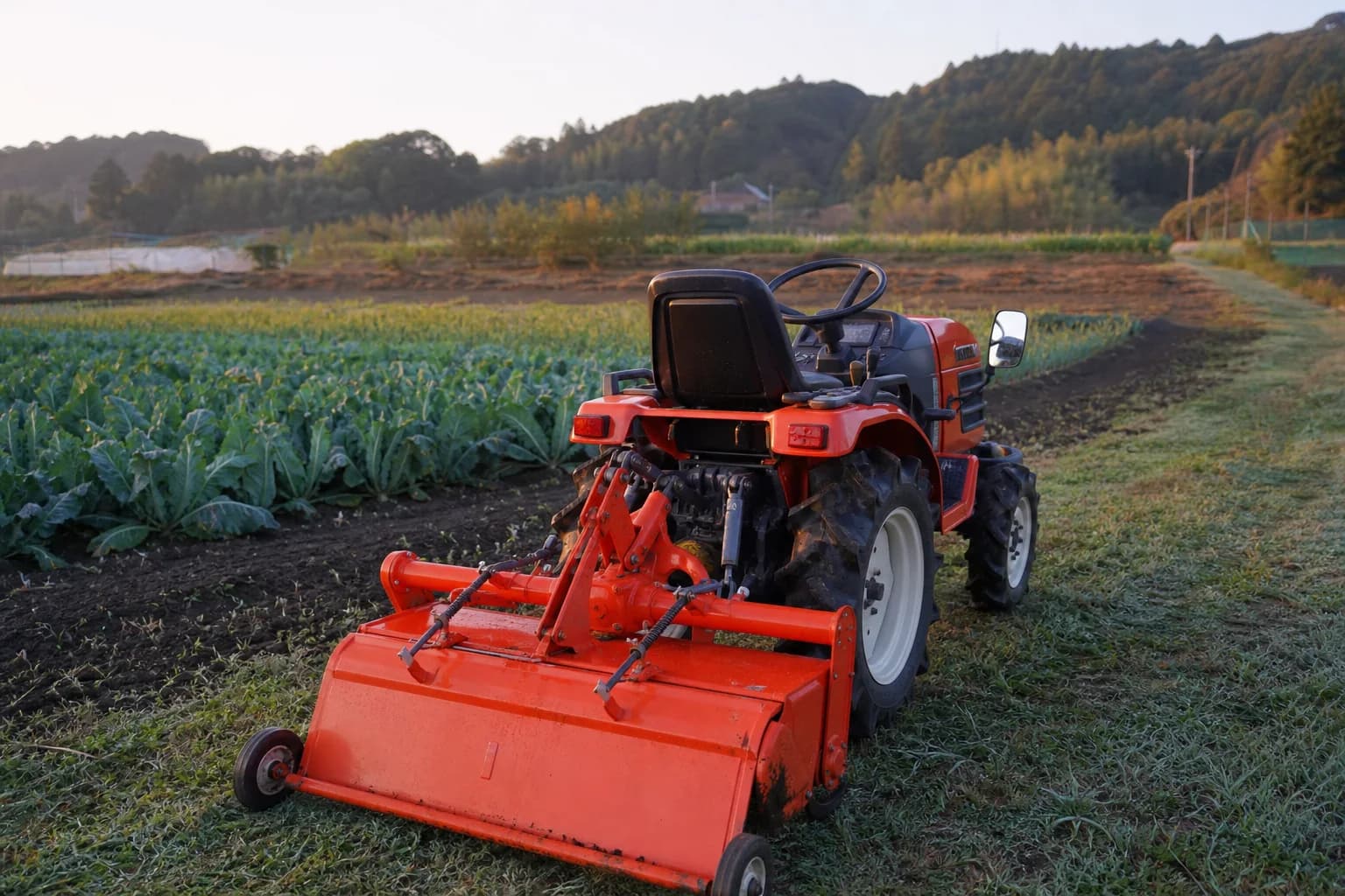 Kompaktný malotraktor s pripojeným náradím stojí na okraji menšieho poľnohospodárskeho pozemku.