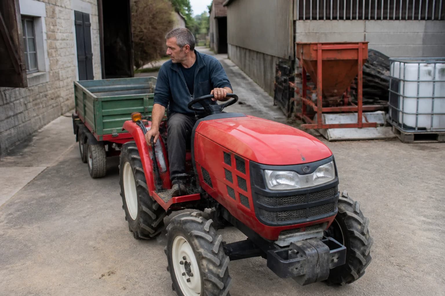 Kompaktný malotraktor na dvore s vodičom pri ovládacej páke a pracovným náradím v pozadí.
